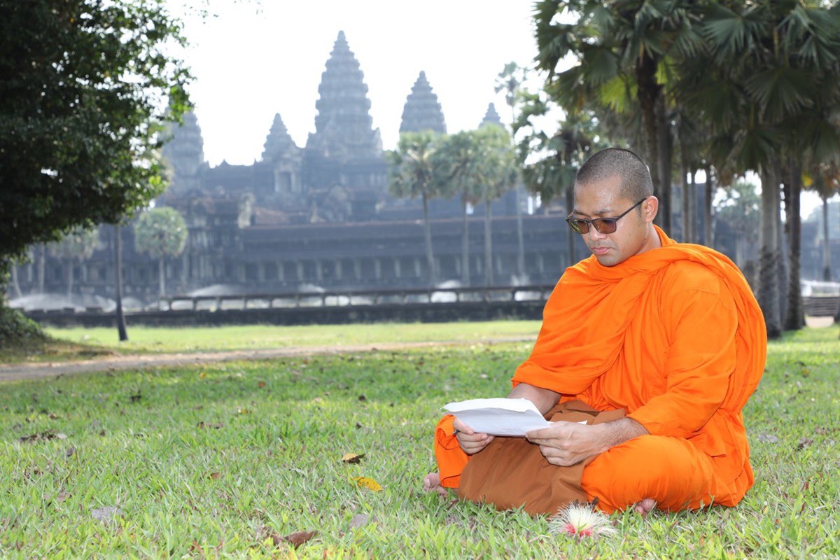 The author Lay Lon is pictured sitting cross-legged on green grass wearing tinted glasses and saffron robes. He is reading from a sheet of paper with the temple complex  Angkor Wat in the background.