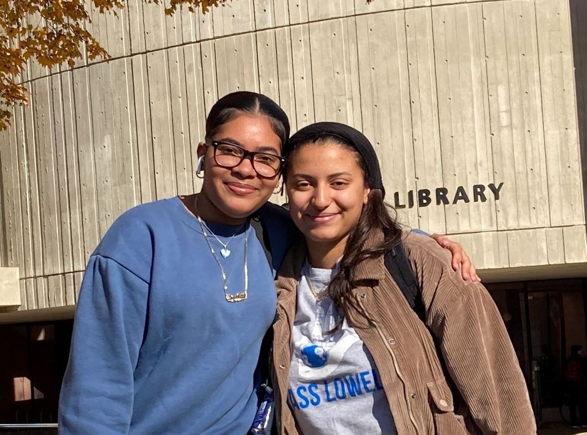 2 students posing outside  together with a building with the word Library on it behind them.