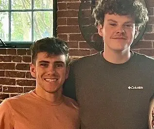Two people in T-shirts pose for a photo in front of a brick wall and window.