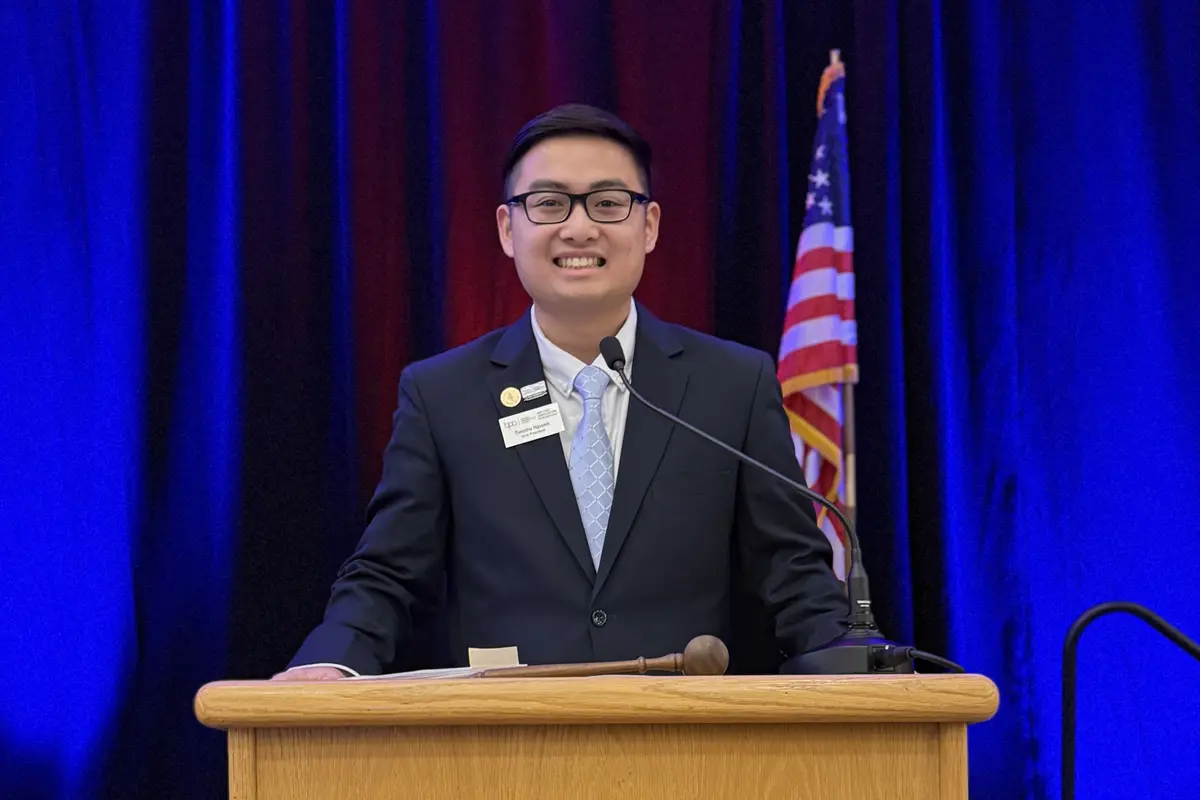 A young man in glasses and a suit poses for a photo at a podium.