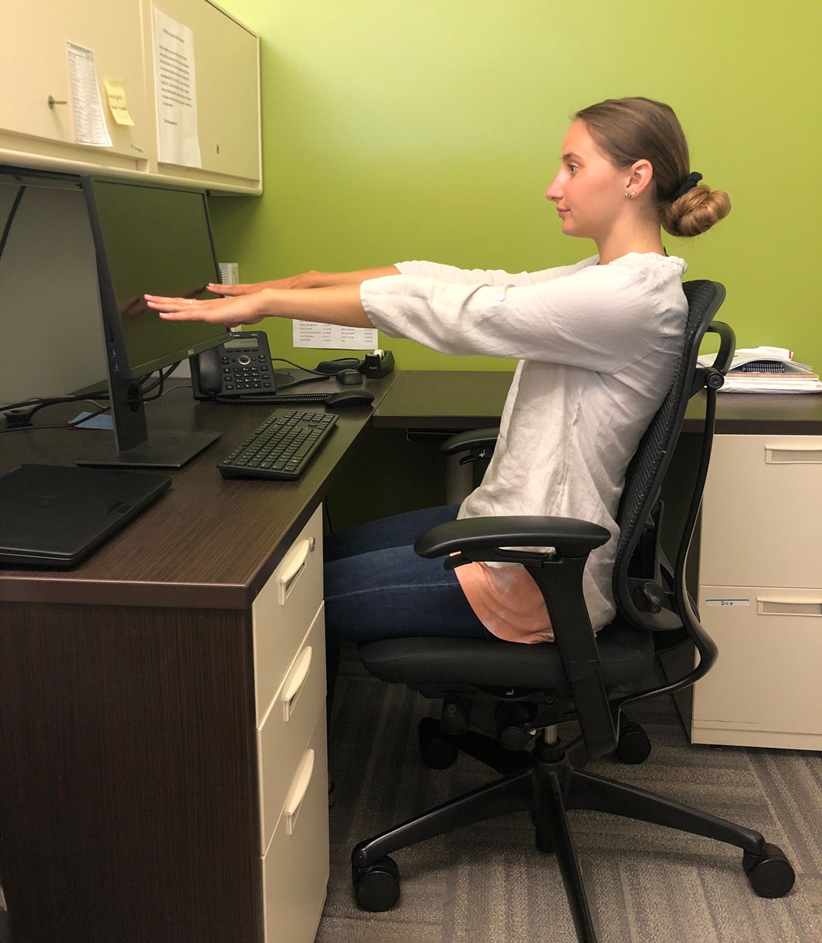 A person sits at her desk with their arms extended in front of them so that the tips of their fingers touch the monitor.