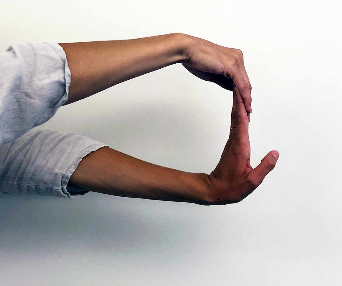 A person extends both arms in front of them. The bottom hand has the palm facing away from them and the fingers pointed towards the ceiling. The top hand is pulling the fingers of the bottom hand towards the body for a stretch.