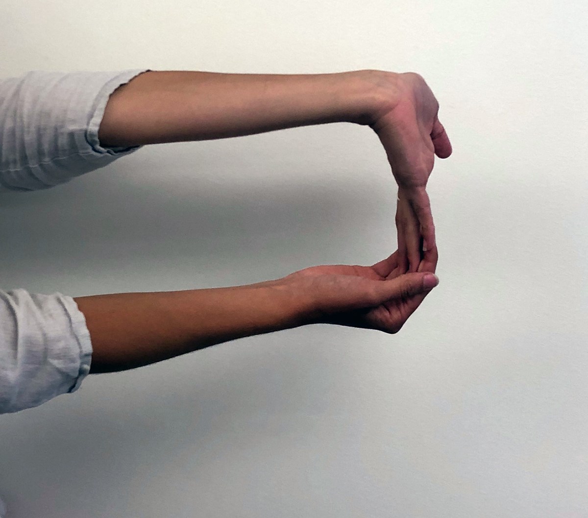 A person extends both arms in front of them. The top hand has the palm facing away from them and the fingers pointed towards the ground. The bottom hand is pulling the fingers of the top hand towards the body for a stretch.