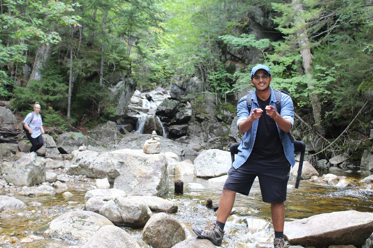 Man wearing backpack standing near stream pointing at camera with smile