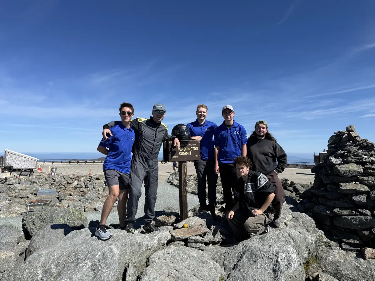 Six young men pose for a photo next to a sign on the summit of a mountain.