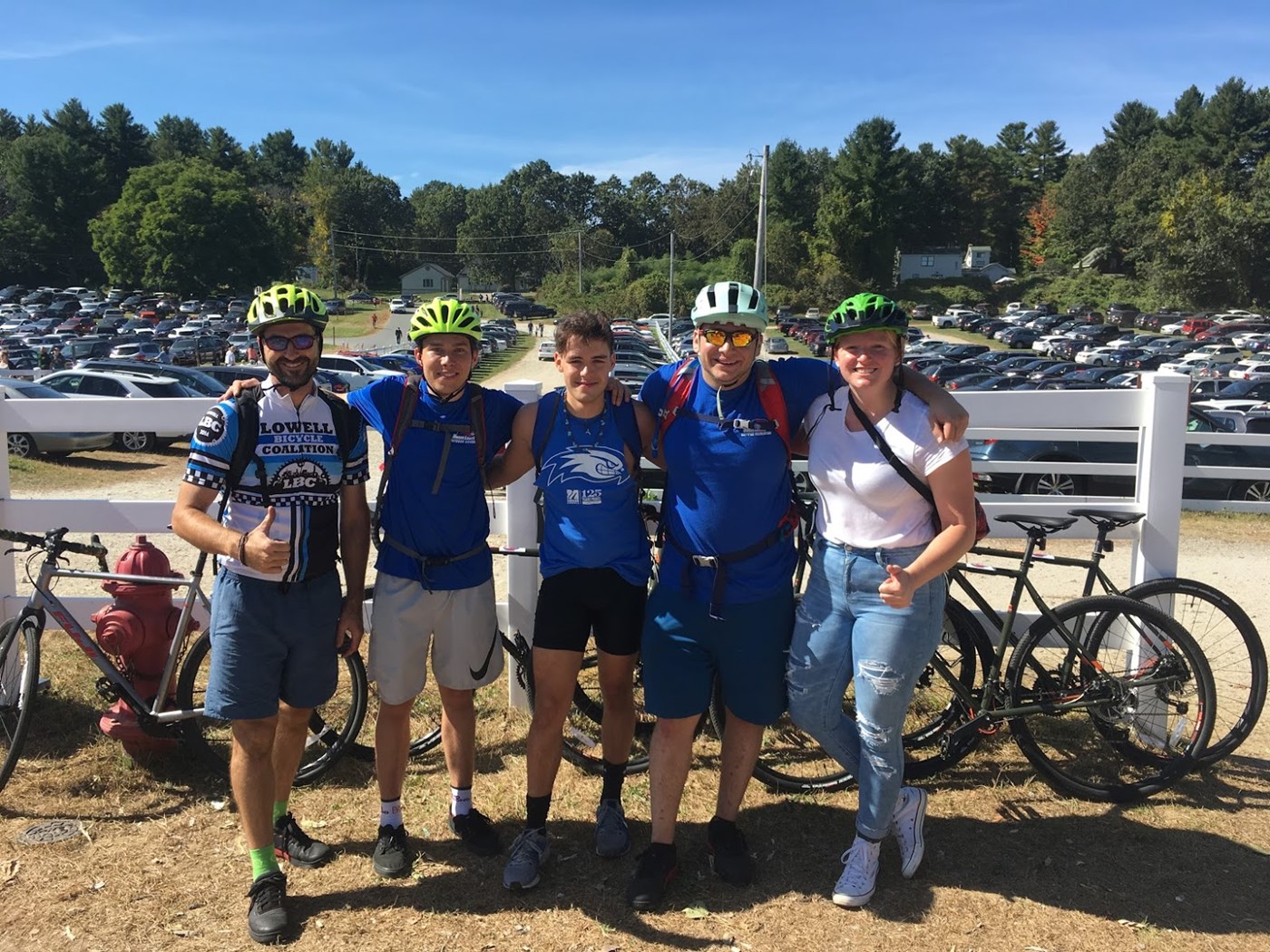 A group of trip participants surrounded by their bikes with arms around one another, thumbs up, and helmets on.