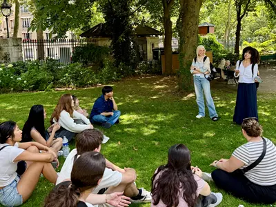 Two young women stand in park and talk to a group of students who are seated in the grass.