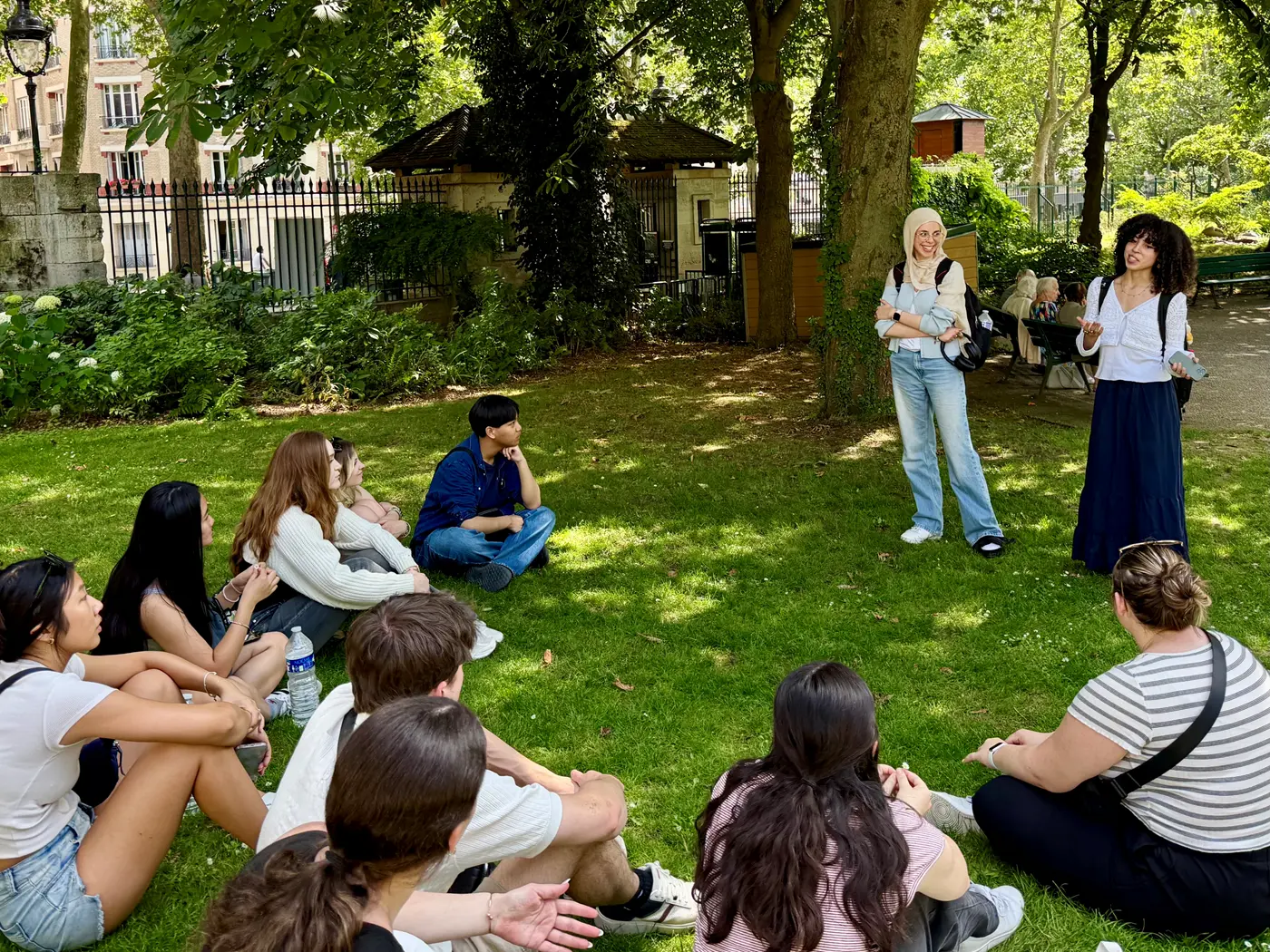 Two young women stand in park and talk to a group of students who are seated in the grass.