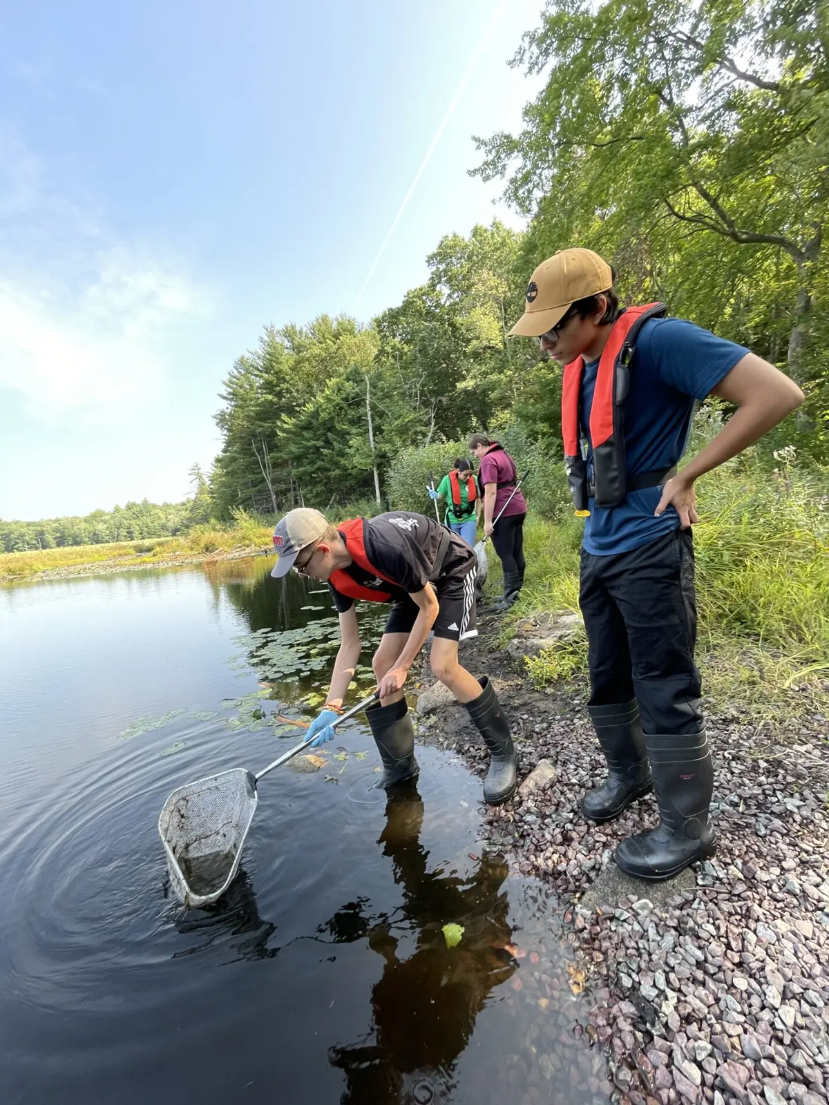 Four students standing on river bank, one scooping something from water with a net