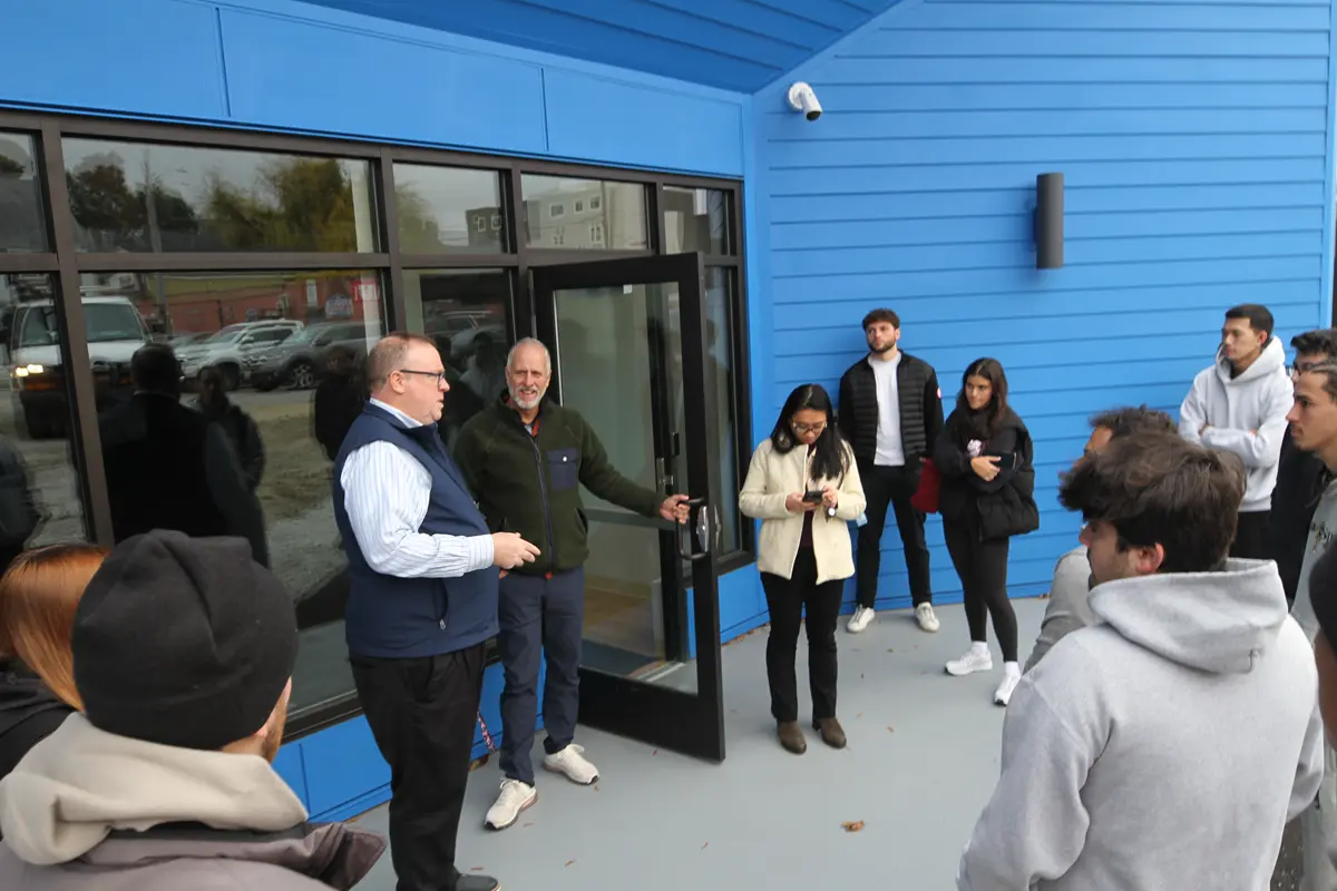 A man in a vest talks to a group of students outside a building while a man holds the door open.