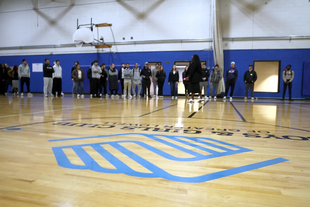 A group of students listen to a woman speak inside a gymnasium.