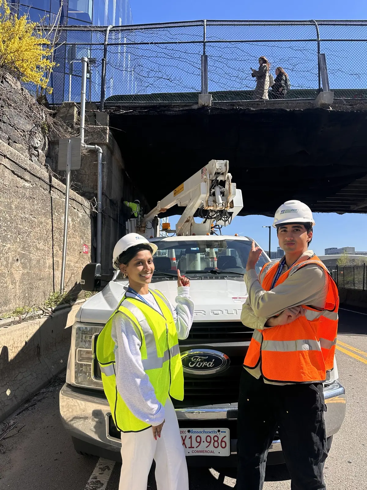 A young woman and man in hard hats and safety vests point to a bridge while standing in front of a truck.