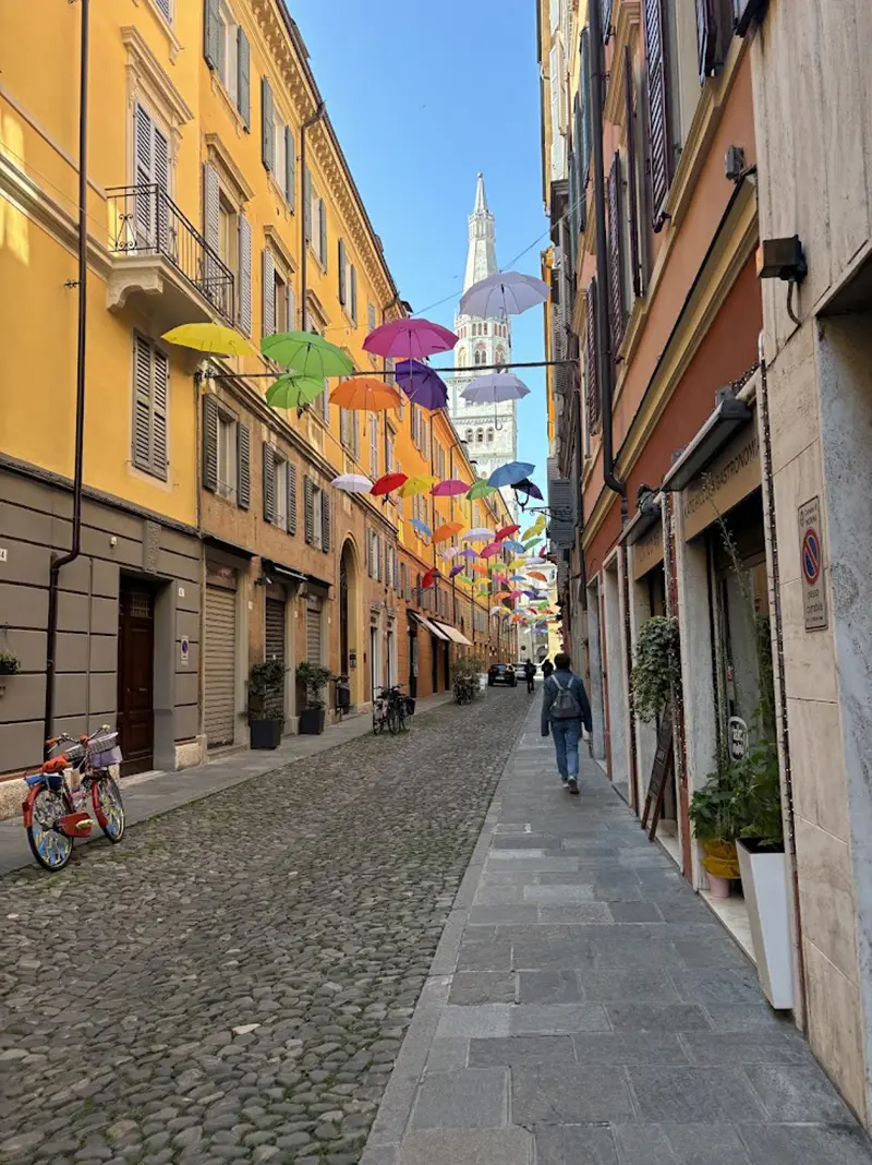 View of Via Cesare Battisti in Modena, with colorful umbrellas suspended overhead and people walking along the street.