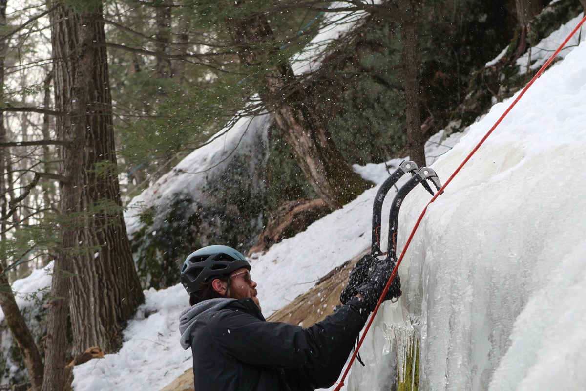 small bits of ice fly as a man in a helmet jams his ice tools into the ice wall while climbing