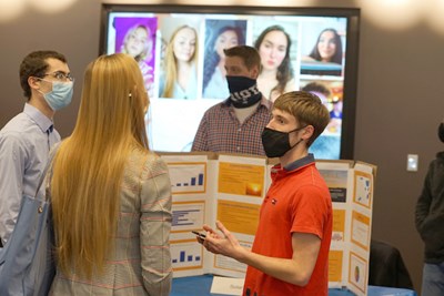 A male student in an orange shirt and face covering explains his project to a woman