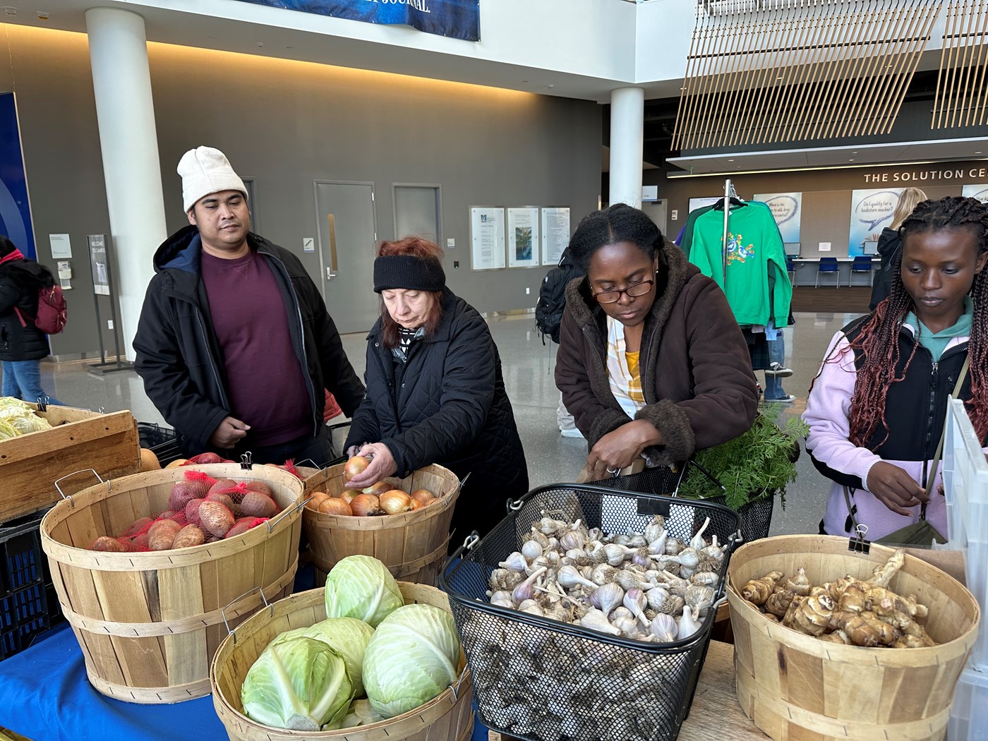 People gather at an indoor farmers market at a table featuring potatoes, onions, cabbage, garlic, and vegetable roots. 