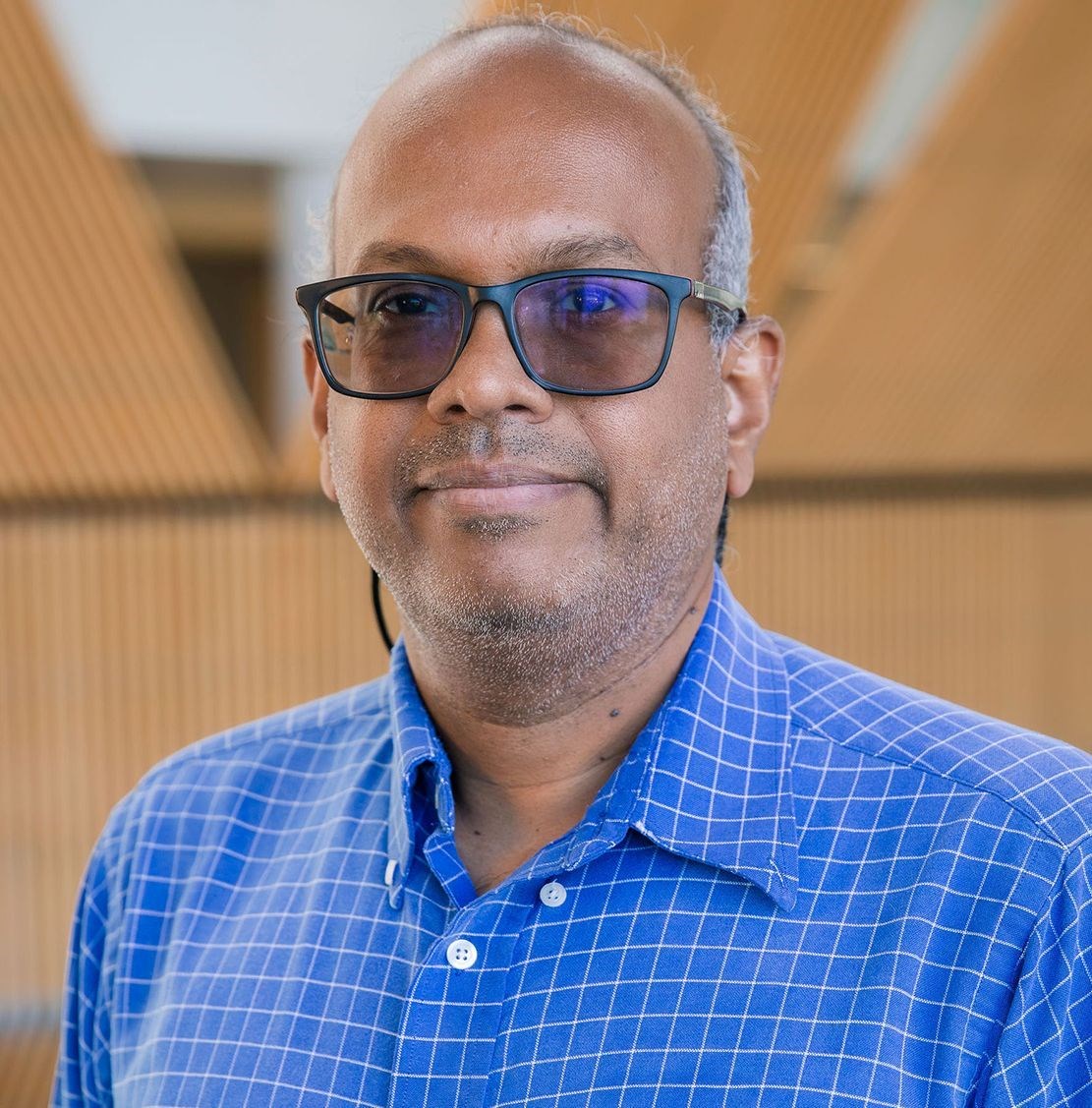 Suri Iyer wearing a blue striped collared shirt and darkened glasses and looking at the camera.