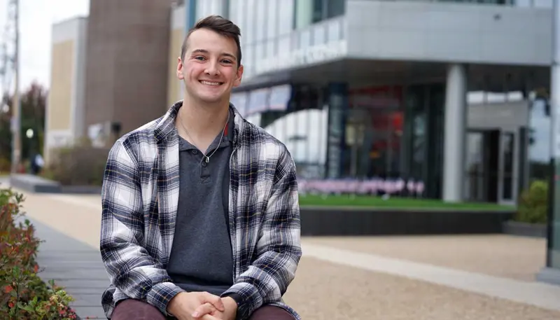 Jacob Villeneuve sits outside with UMass Lowell's University Crossing building in the background.