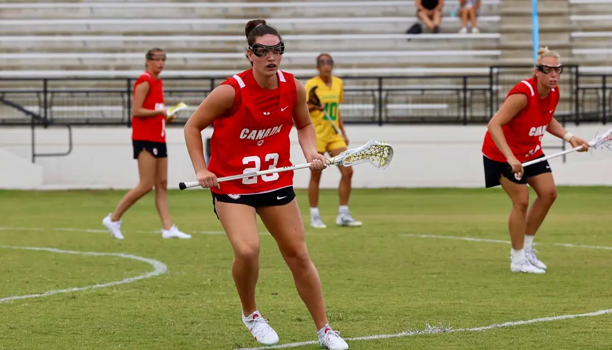 Jillian Goldie holding a lacrosse stick on the playing field with players from the Canadian Women's Lacrosse National Team.
