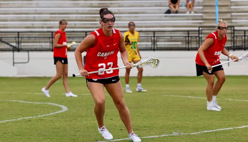 Jillian Goldie holding a lacrosse stick on the playing field with players from the Canadian Women's Lacrosse National Team.