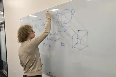 A person in a tan shirt writes on a dry erase board in a college classroom.