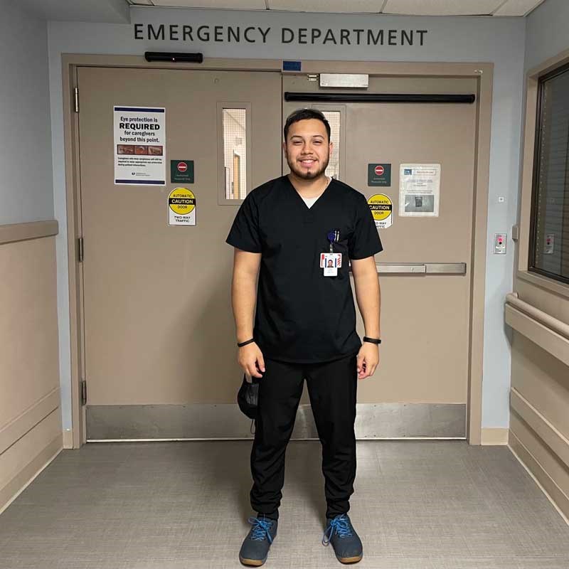 Jose Archila Quezada is wearing scrubs and standing outside the doors to an emergency department in a hospital.
