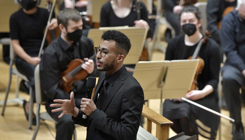 Music education student Josh Santana stands holding a microphone with members of the Me2/Orchestra in the background.
