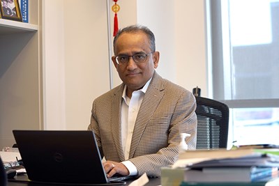 A man in glasses and a blazer poses for a photo while working on a laptop at his desk