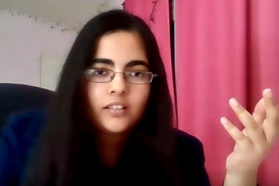 A female student speaks on webcam while waving her left hand in front of a red curtain