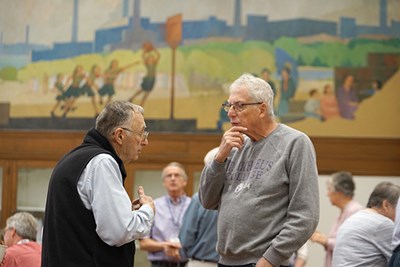 Two men have a conversation while standing in a room with a mural on the wall behind them