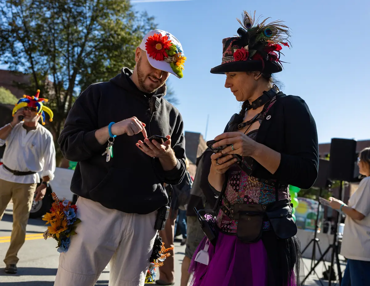 A young man wearing a cap covered in flowers looks at his phone while talking to a woman wearing a hat with feathers outdoors.