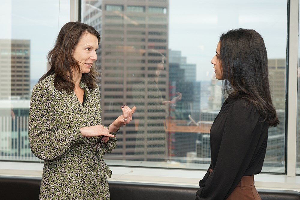Two women talk in front of a window in an office building.