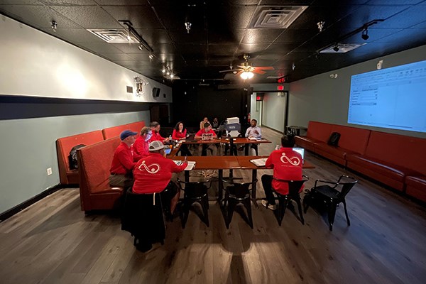 A half dozen people in red shirts sit around a table and look at a projection screen during a meeting