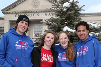Four students in UMass Lowell sweatshirts in front of snowy Cumnock Hall