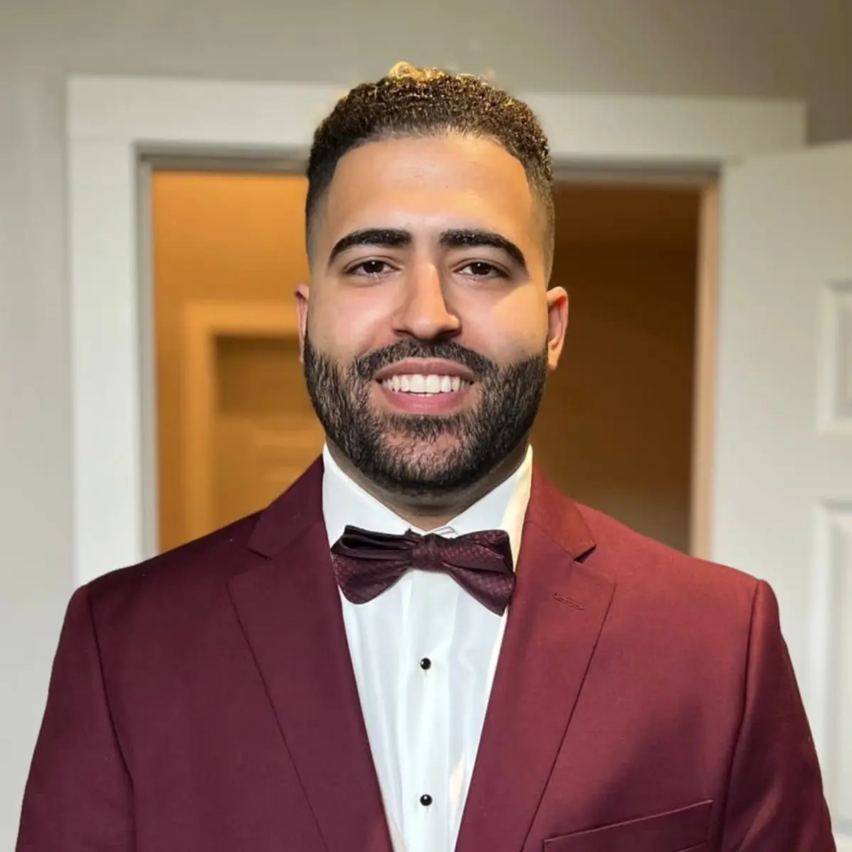 A young man in a maroon suit and bowtie poses for a photo in a room.