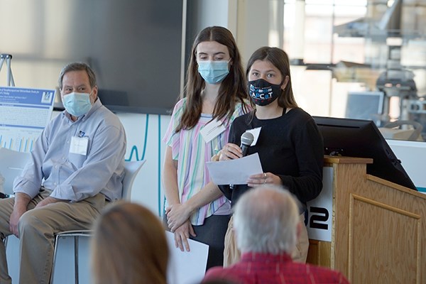 A female student wearing a masks holds a microphone and paper while speaking, while a man and a woman look on