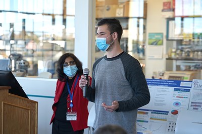 A male student wearing a mask holds a microphone while talking as a woman in glasses and a red jacket looks on