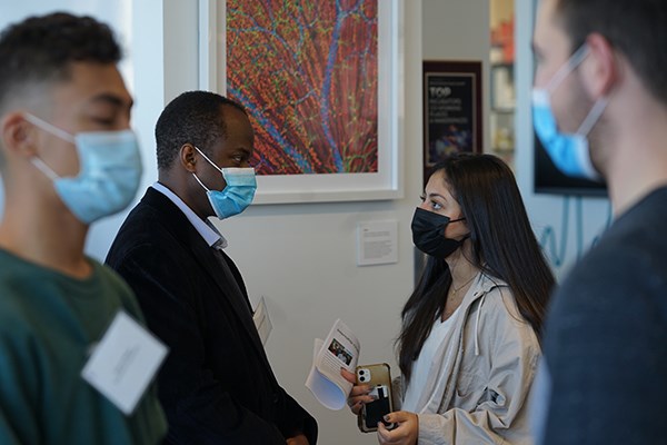 A masked woman with dark hair and holding a phone and papers speaks with a man who is wearing a mask