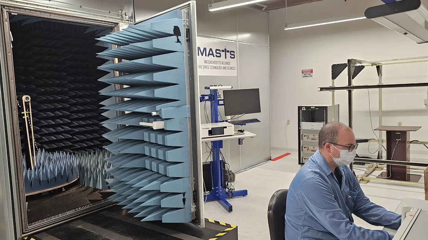 Man wearing a mask works at a laptop in front of metal door with blue spikes on it and MASTS sign on wall