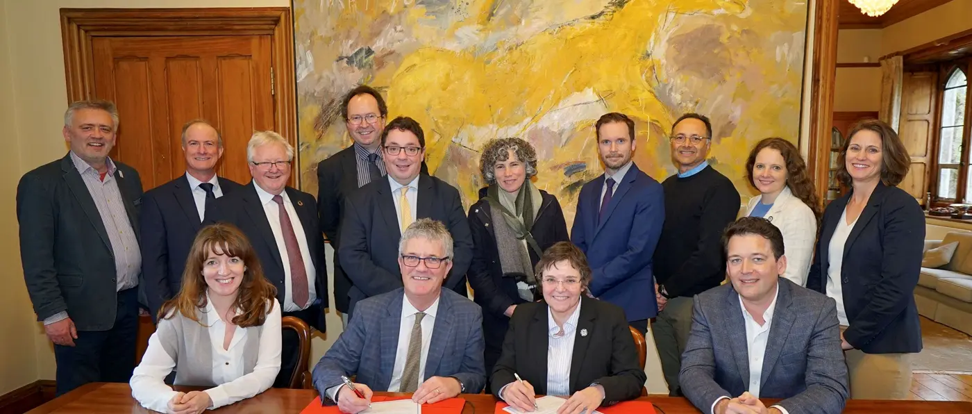Four people sitting and 10 people standing at a conference room table where a 2 people are signing documents