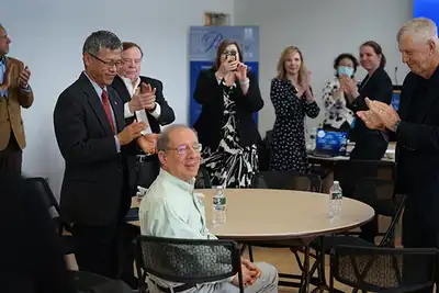 People stand and applaud for a man who is sitting at a table