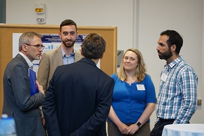 Four men and a woman talk to each other in front of a poster presentation