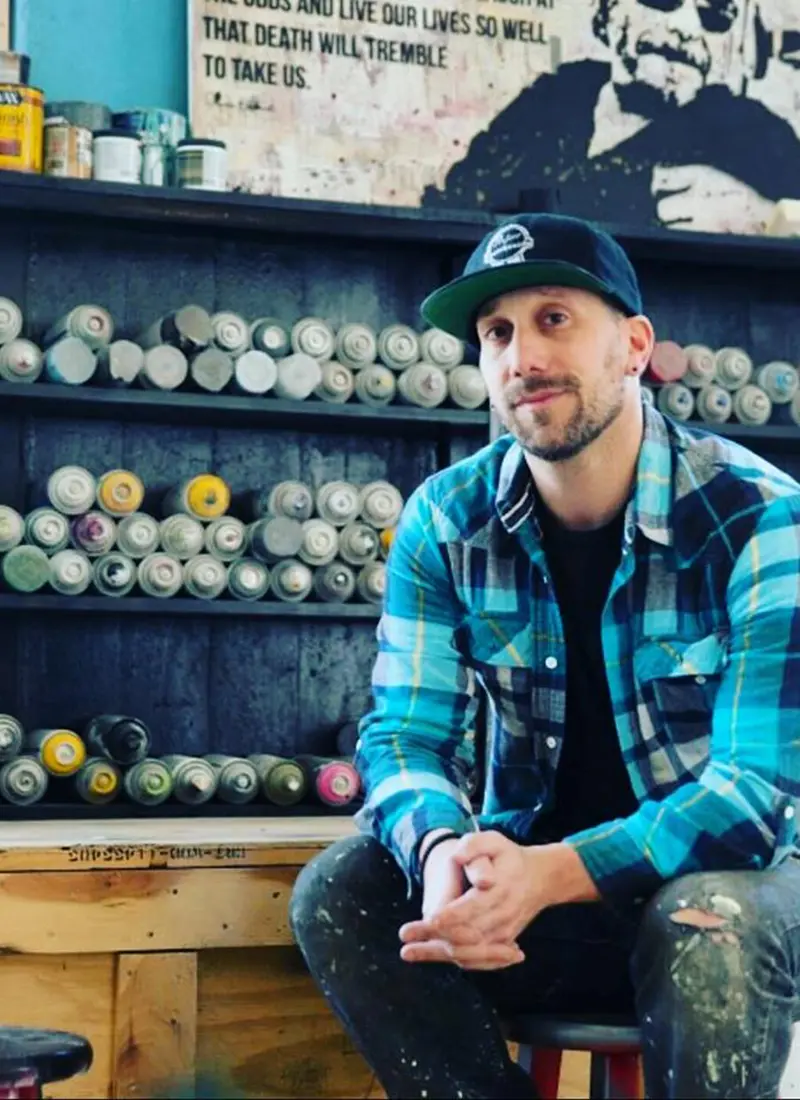 Markus Sebastiano in an art studio sitting in front of rows of spray paint cans