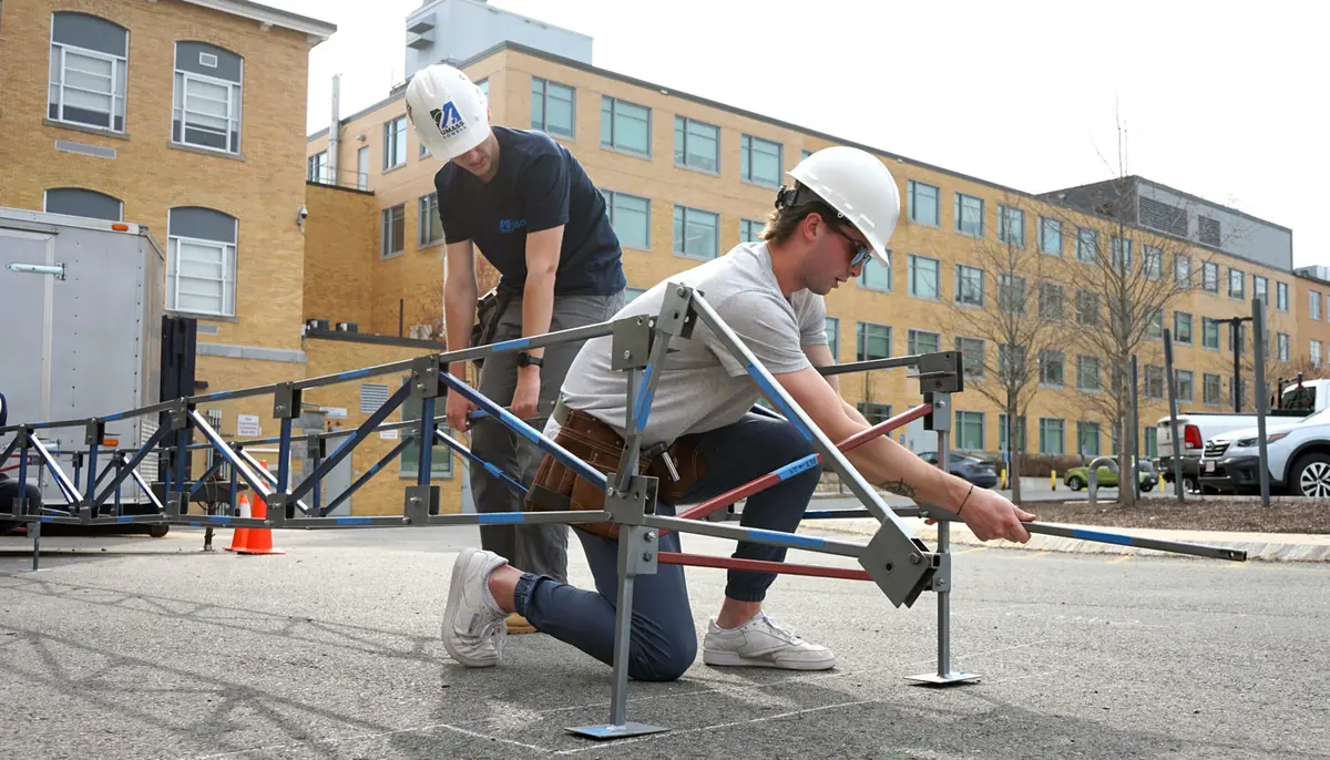 Matt Paxton works outdoors with another person building a steel bridge.