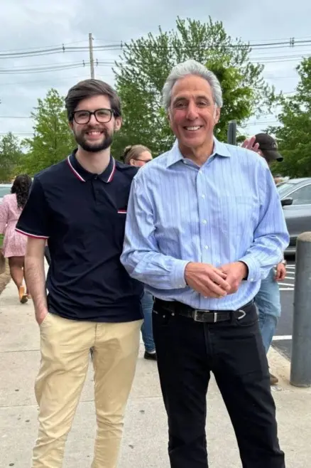 A college student with a beard and glasses poses for a photo with a man in a blue button down shirt outside.