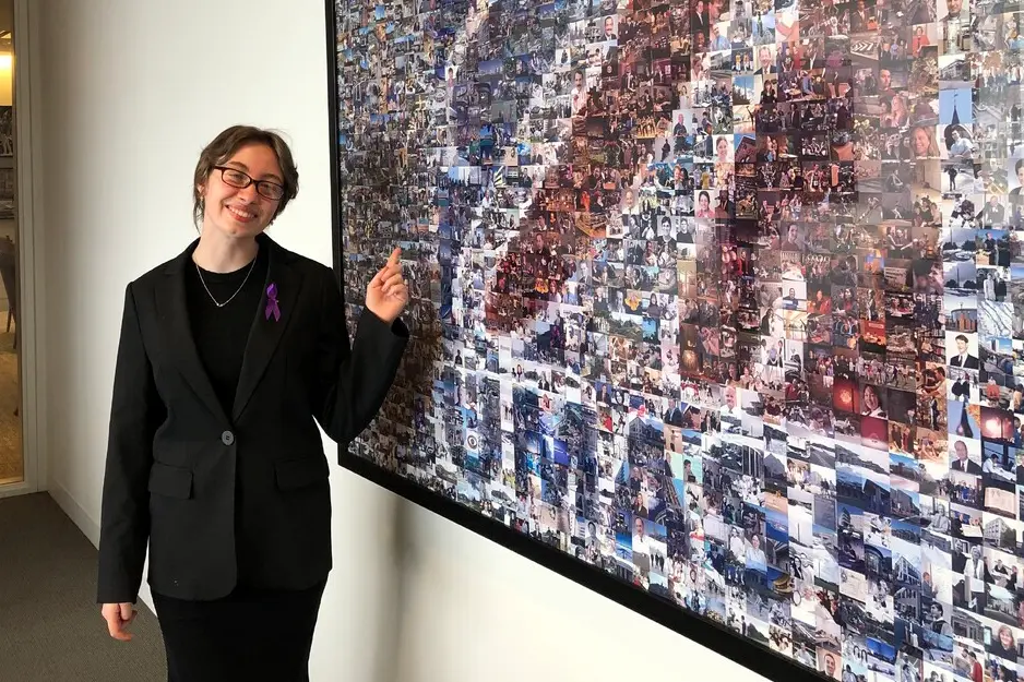 A young woman in a dark suit and glasses poses for a photo next to a photo collage that makes the UMass colllege logo.