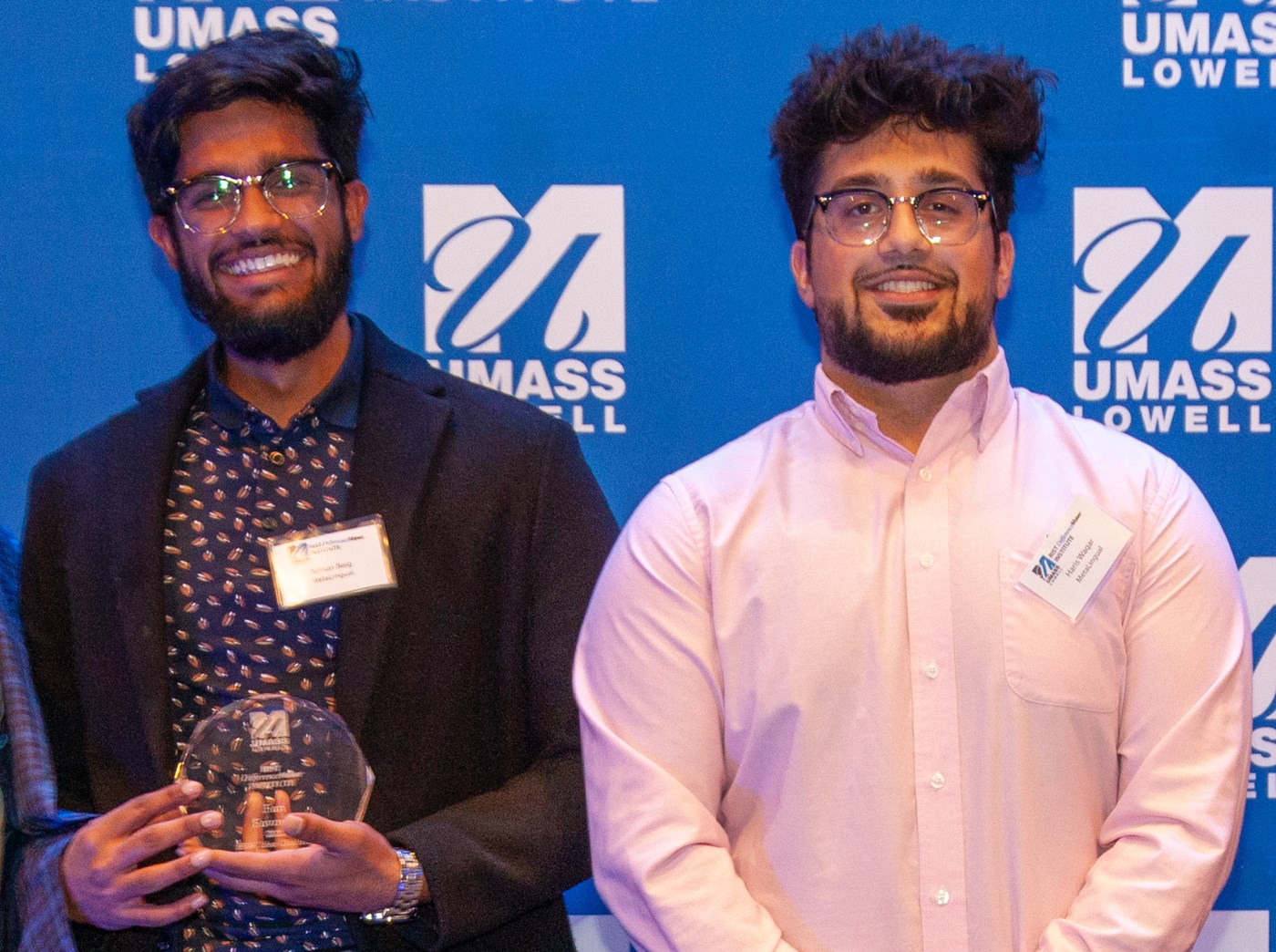 2 male students from the MetaLingual team holding an award and posing against a blue UMass Lowell backdrop.
