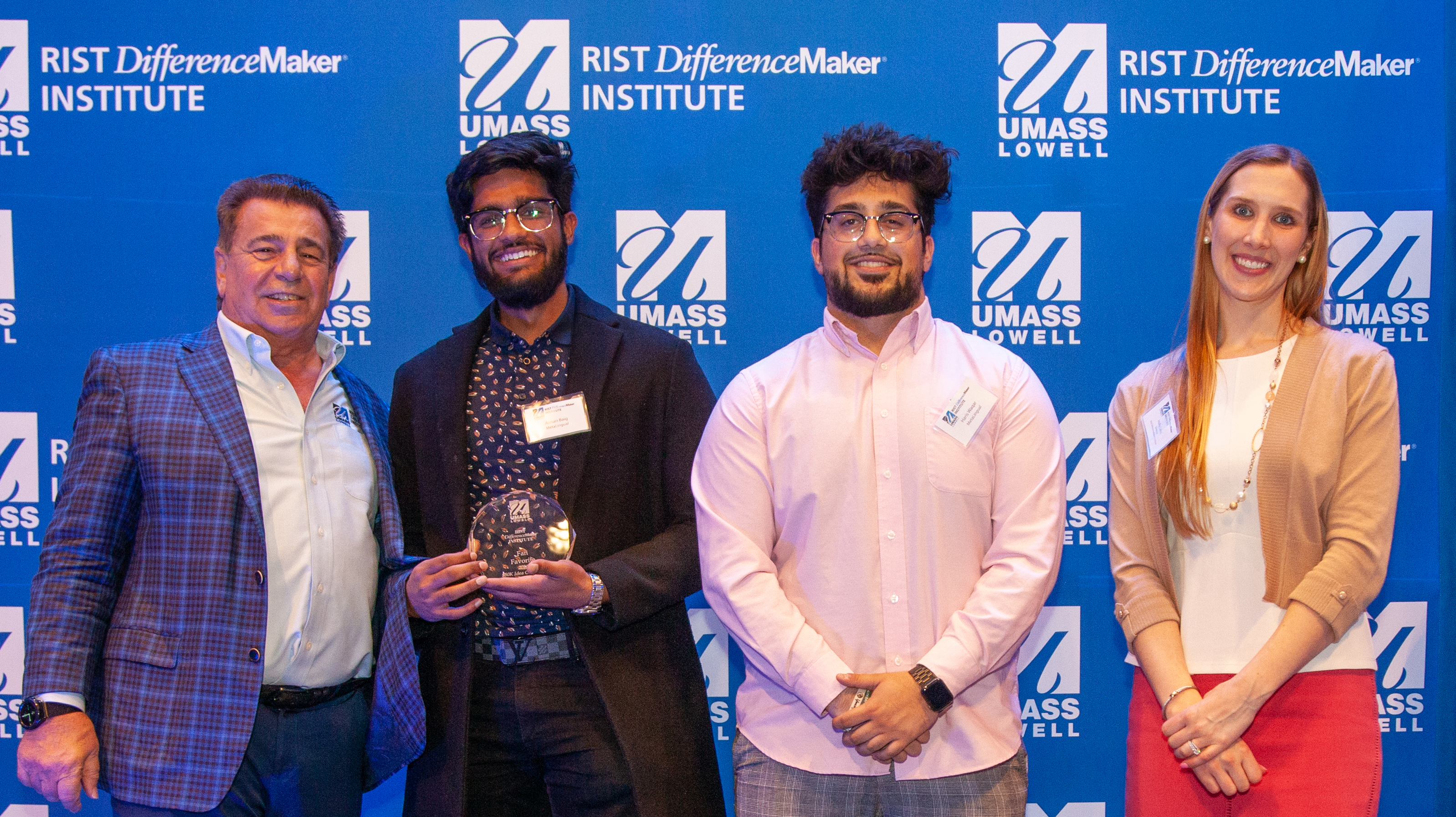 2 male students from the MetaLingual team holding an award and posing with Brian Rist and Holly Lalos from Difference makers against a blue UMass Lowell backdrop.