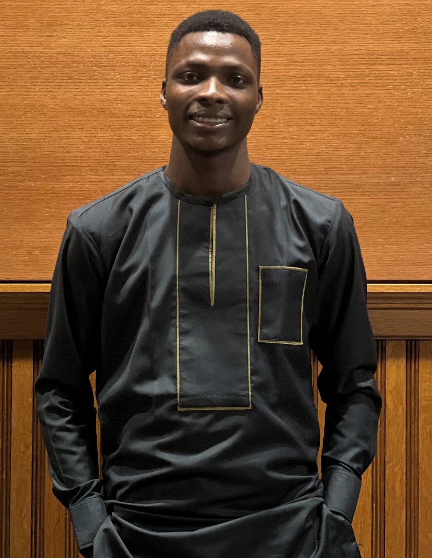 Michael Olufemi wearing a black and gold shirt standing in front of a wood background.