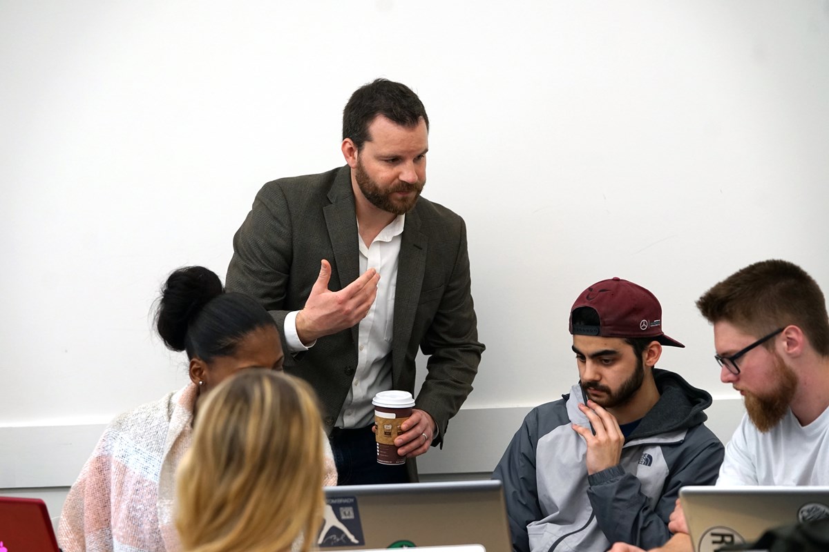 Assistant Professor Michael Obal talks to a group of business students on their laptops.